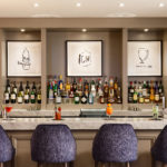 The bar at mercure gloucester bowden hall hotel with bottles lined up on the shelf behind and blue bar stools neatly arranged in front